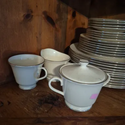 Sale Photo Thumbnail #4: Vintage white porcelain china service set with silver trim, featuring a covered sugar bowl, creamer pitcher, teacup, and stacks of plates.