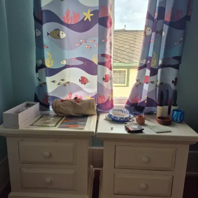 Sale Photo Thumbnail #96: Pair of off-white two-drawer nightstands featuring bun feet and round knobs, shown beneath ocean-themed fish curtains.