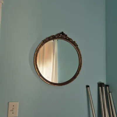 Sale Photo Thumbnail #113: Vintage round wall mirror with an ornate gold-toned ornate crest and frame, mounted on a light blue wall.