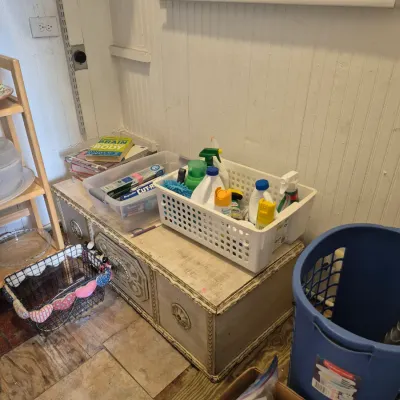 Sale Photo Thumbnail #26: Vintage painted wooden storage bench with decorative molded panel detailing, holding plastic organizer baskets with household cleaning supplies, alongside a wire basket and blue laundry hamper.