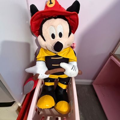 Large Disney Mickey Mouse firefighter plush toy, seated in a small pink plastic cart, dressed in a red firefighter helmet and yellow coat...