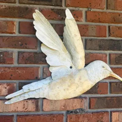 Off-white carved wooden hummingbird wall sculpture featuring detailed feather textures, mounted on a red brick wall.