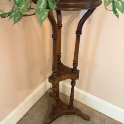 Traditional dark wood plant stand featuring turned support legs, two lower triangular tiers, and a rounded top surface, displayed with a...