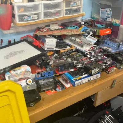 Large assortment of packaged die-cast toy cars and collectibles, including Hot Wheels and Monster Jam brands, cluttering a workbench...
