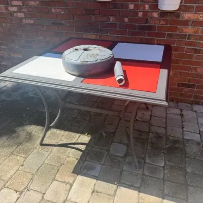 Outdoor patio dining table featuring a metal frame with a red and white tiled top, topped with a heavy umbrella base weight.