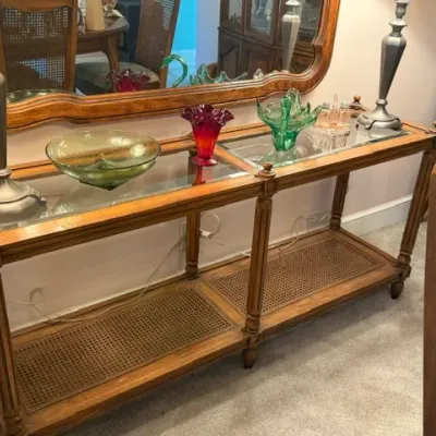 French Provincial style console table featuring glass insert tops, a lower woven cane shelf, fluted turned legs, and displayed decorative...