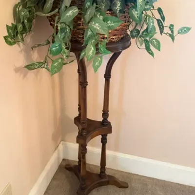 Tall vintage wooden plant stand featuring turned spindle legs and a three-legged base, holding a faux potted plant in a woven basket.