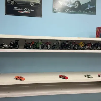 Collection of scale model motorcycles and die-cast cars displayed on white wooden wall shelves below classic car metal signs.