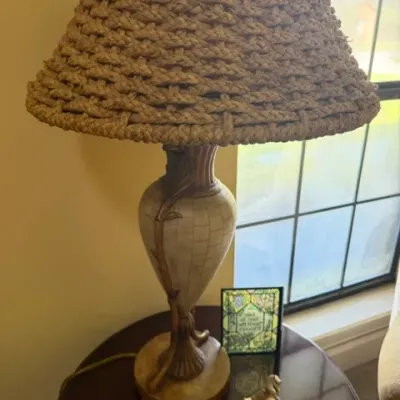 Rustic table lamp featuring a ceramic urn-style base with branch accents and a heavy woven rope lampshade, shown resting on a dark wood...