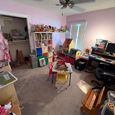 Child's bedroom filled with toys, plush animals, dollhouses, a small play table with chairs, a white cube organizer shelf, and...