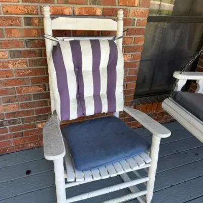 White painted wooden porch rocking chair featuring turned posts, a slatted seat, and cushioned seat and back pads.