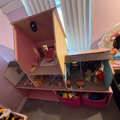 Large wooden two-story dollhouse furnished with miniature furniture and figures, resting atop a pink storage unit with fabric bins.