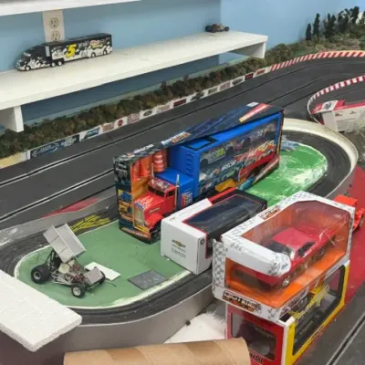 Collection of die-cast model vehicles and toy NASCAR hauler trucks displayed on a large slot car race track table.