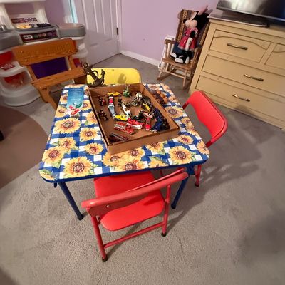 Children's activity room featuring a sunflower-patterned card table set with red folding chairs, a box of toy cars, a play kitchen,...