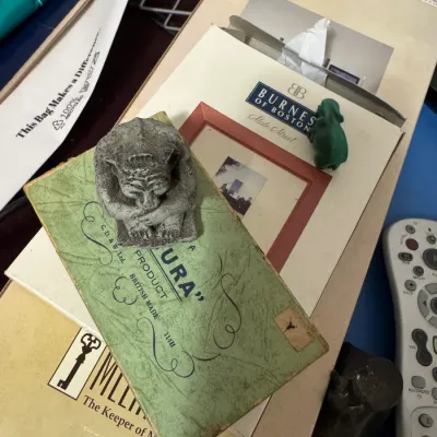 Sale Photo Thumbnail #56: Small gray concrete gargoyle figurine resting on top of a vintage British-made paper box alongside a green figurine, boxed Burnes of Boston frame, and a television remote.