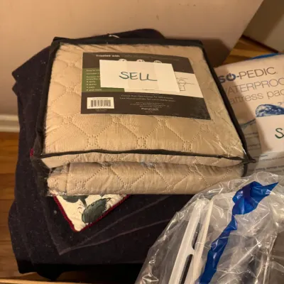 Sale Photo Thumbnail #94: Quilted beige furniture protector cover in original clear zippered packaging marked 'SELL', resting atop other household items.