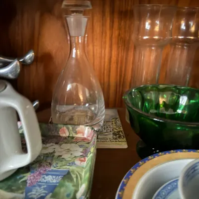 Sale Photo Thumbnail #116: Collection of glassware and tableware, including a clear decanter, green glass bowl, floral printed coasters, and ceramic dishes displayed in a wooden cabinet.