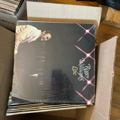 Sale Photo Thumbnail #159: A cardboard box filled with vinyl LP records, featuring a shrink-wrapped copy of 'Roger Williams Live' on top.