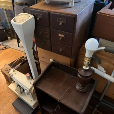 Sale Photo Thumbnail #140: Collection of vintage household items including a multi-drawer wooden card catalog cabinet, a small dark wood nightstand, a vintage sewing machine, and a floor scale.