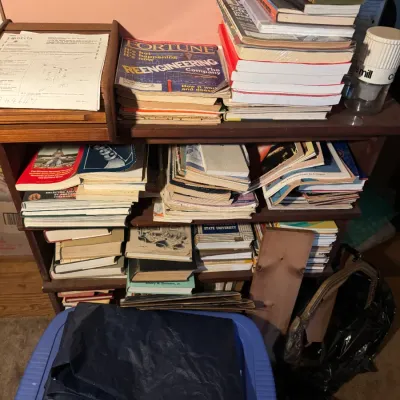 Sale Photo Thumbnail #78: Wooden shelving unit packed with stacks of vintage books, manuals, and magazines, including Fortune magazine. A folded blue tarp sits on top of a plastic bin in the foreground.