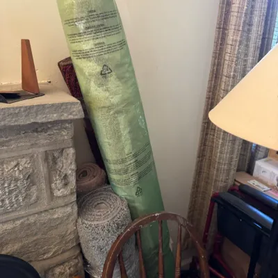 Sale Photo Thumbnail #155: Rolled carpet runner enclosed in green plastic packaging, stored upright alongside additional rolled rugs and household items.