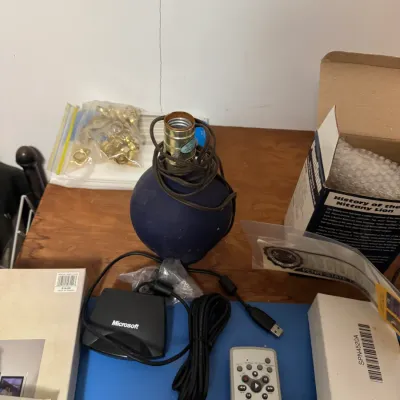 Sale Photo Thumbnail #55: Collection of tabletop items including a round blue ceramic lamp base with coiled cord, a Microsoft USB receiver, and various remotes and boxed goods.