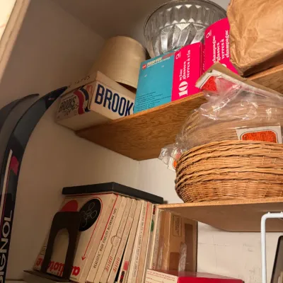 Sale Photo Thumbnail #81: Shelves containing a collection of vintage items including reel-to-reel recording tapes, a Brooks shoe box, a Pana-Vue slide viewer, woven baskets, glassware in original boxes, and a Rossignol ski.