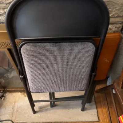 Sale Photo Thumbnail #156: Folded black metal chair featuring a grey textured fabric padded seat, leaning against a stone wall.