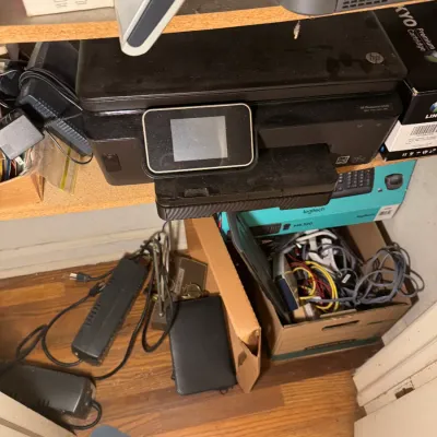 Sale Photo Thumbnail #166: Black HP Photosmart all-in-one printer sitting on a shelf alongside various electronic accessories, cables, and power supplies.