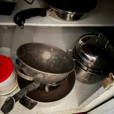 Sale Photo Thumbnail #66: Assorted stainless steel and aluminum cookware, including a metal colander, a skillet, and nested pots inside a white cabinet shelf.