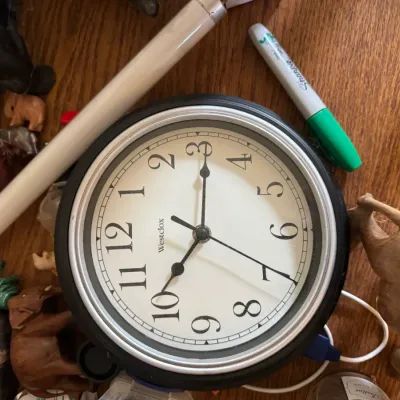 Sale Photo Thumbnail #126: Round Westclox wall clock featuring a black frame and silver bezel, displayed on a wooden surface surrounded by small carved animal figurines.