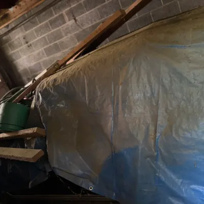 Sale Photo Thumbnail #169: Large item covered by a dusty blue plastic tarp in a garage, flanked by miscellaneous wood lumber and a green bucket.