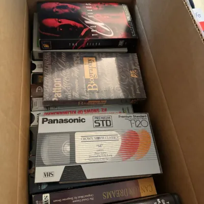 Sale Photo Thumbnail #147: Cardboard box packed with various vintage VHS tapes, including titles like The X-Files, Biography series, and art documentaries.
