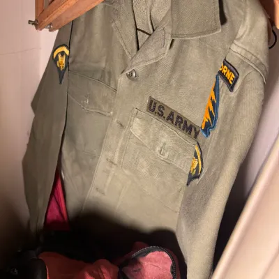 Sale Photo Thumbnail #11: Vintage US Army olive drab utility shirt featuring an Airborne insignia patch and military unit shoulder patches, hanging on a wooden hanger.