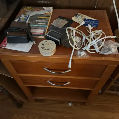 Sale Photo Thumbnail #134: Two-drawer wooden nightstand topped with various household items, power cords, and small electronics.