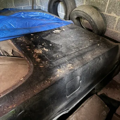 Sale Photo Thumbnail #164: Dusty vintage black car body partially covered by a blue tarp, stored in a cinderblock garage with spare tires behind it.
