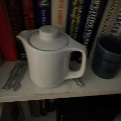 Sale Photo Thumbnail #106: White ceramic teapot resting on a bookshelf alongside a dark ceramic mug and various hardcover books.