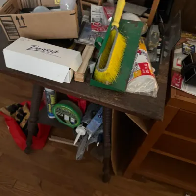 Sale Photo Thumbnail #135: Assorted household supply items, including a long yellow-bristled brush, light bulbs in a cardboard box, tape, and sealant, sitting atop a wooden table.