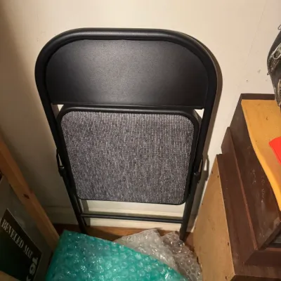 Sale Photo Thumbnail #86: Folded black metal frame chair featuring a textured grey upholstered seat cushion, resting against a wall next to packing materials.