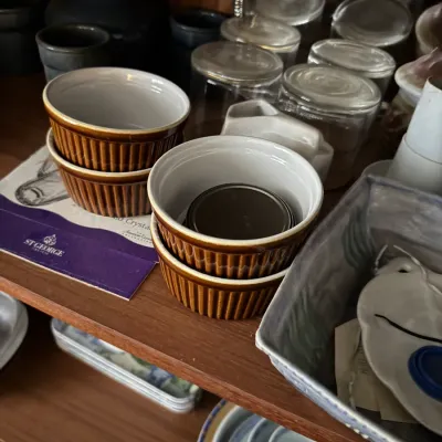 Sale Photo Thumbnail #107: Collection of brown ribbed ceramic ramekins displayed alongside clear glassware and additional kitchenware on wooden shelves.
