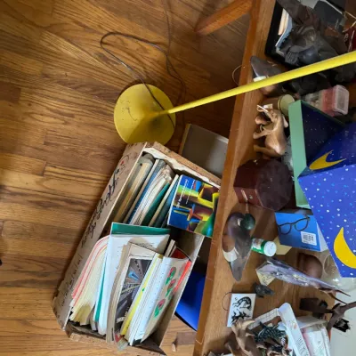 Sale Photo Thumbnail #161: Collection of eclectic home goods, including wooden carvings, books, and ephemera stored in crates and arranged on a low table alongside a yellow floor lamp.