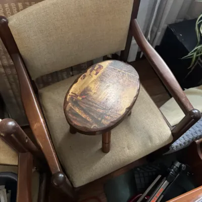 Sale Photo Thumbnail #14: Vintage small oval wooden footstool with a decoupaged landscape scene, placed on a mid-century wooden armchair with beige fabric upholstery.