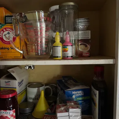 Sale Photo Thumbnail #61: Kitchen cabinet shelf filled with miscellaneous household items, including Pyrex measuring cups, paint bottles, boxes of matches, and various packaged foods.