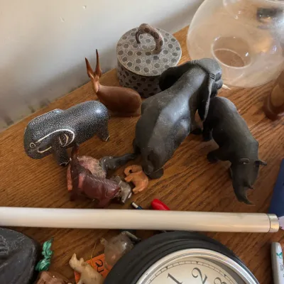 Sale Photo Thumbnail #131: Collection of animal figurines, including carved soapstone and wooden elephants, a rhinoceros, and an antelope, displayed alongside a round Westclox wall clock.