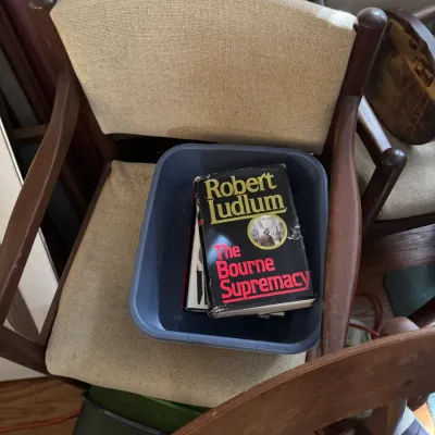 Sale Photo Thumbnail #13: Hardcover copy of Robert Ludlum's "The Bourne Supremacy" sitting inside a plastic bin resting on a mid-century modern style wooden armchair with beige fabric upholstery.