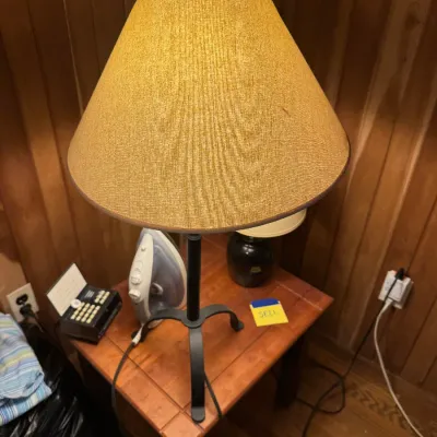 Sale Photo Thumbnail #87: Black wrought iron tripod table lamp with a wide textured fabric shade, resting on a small wooden side table.