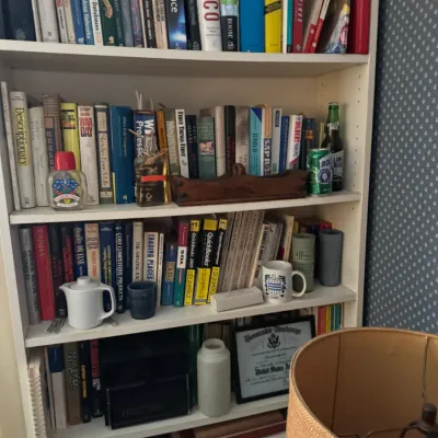 Sale Photo Thumbnail #102: White wooden five-shelf bookcase filled with a varied collection of vintage books, decoratives, teapots, and framed documents.