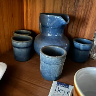 Sale Photo Thumbnail #15: Blue ceramic beverage set including a pitcher and four matching tumblers, displayed on a wooden shelf.