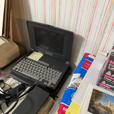 Sale Photo Thumbnail #70: Vintage dark gray laptop computer, likely from the early 1990s, surrounded by office supplies and power cords.