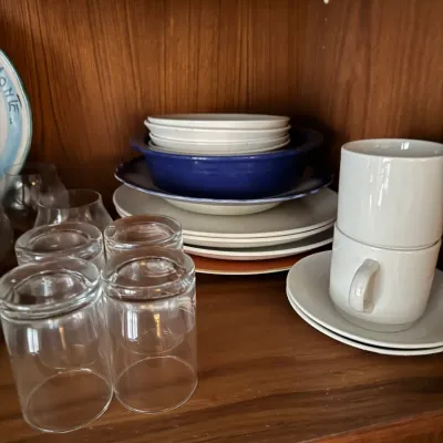 Sale Photo Thumbnail #109: Collection of assorted mid-century modern glassware and ceramic tableware, including clear drinking glasses, white ceramic coffee mugs, and stacked serving plates inside a wood cabinet.